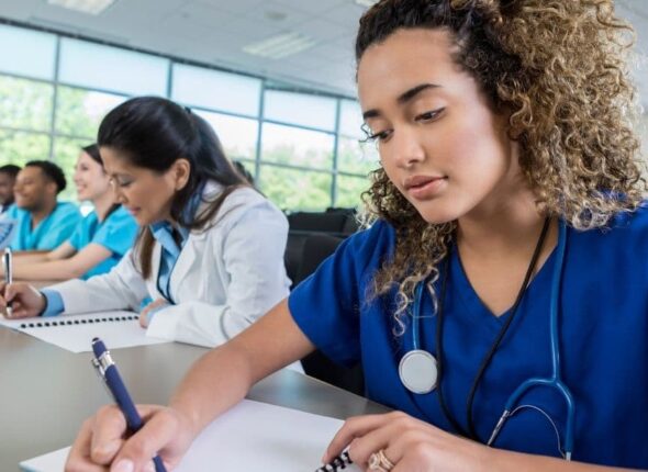 A group of people in medical scrubs and lab coats are seated in a classroom, taking notes attentively.