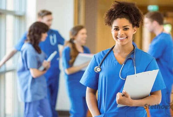 A healthcare professional in blue scrubs holds a clipboard, standing in a hallway with colleagues in the background.