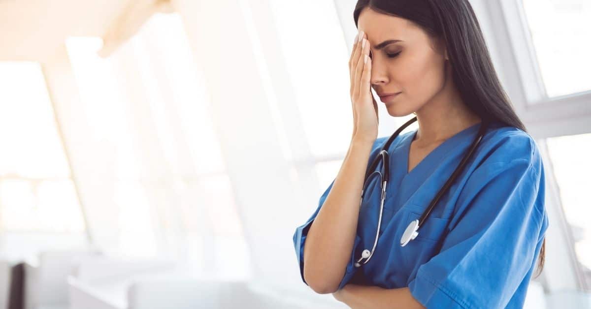 A healthcare professional in blue scrubs appears stressed or fatigued, holding her forehead in a gesture of concern or exhaustion, reflecting common mistakes new CNAs make.