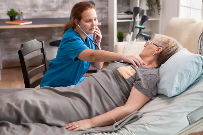 A nurse in blue scrubs uses a stethoscope to examine an older woman lying in bed, covered with a gray blanket.