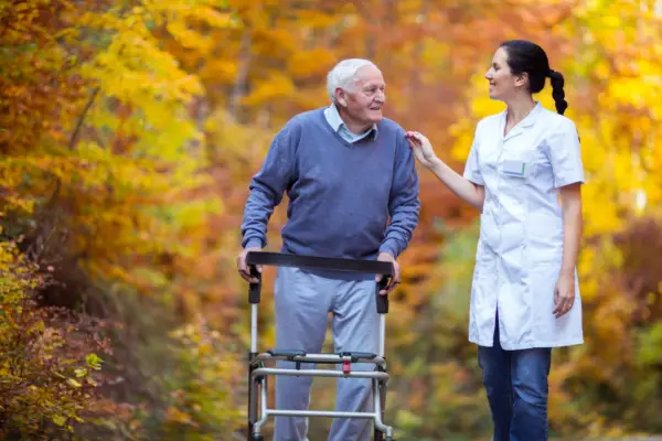 An elderly man using a walker is accompanied by a caregiver in a white uniform. They are walking on a path surrounded by colorful autumn foliage.