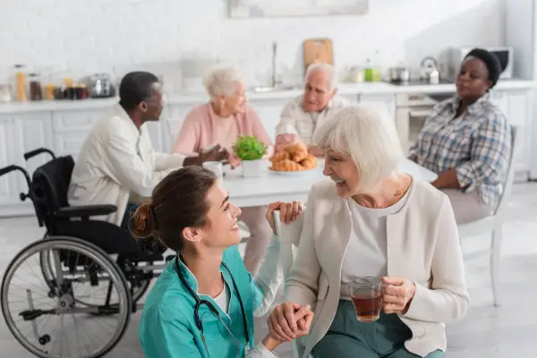 Caregiver kneeling and smiling at an older woman holding tea. Three other seniors and a caregiver are seated at a table in the background.