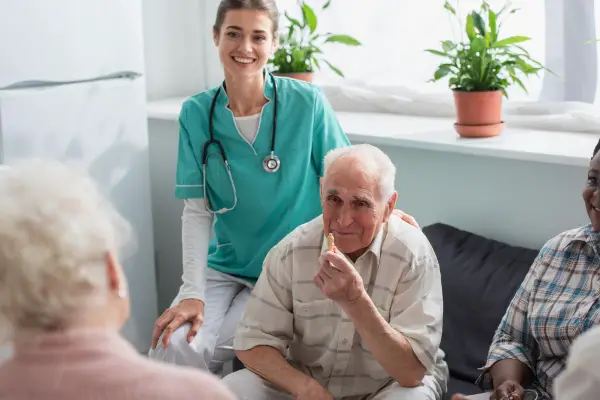 A nurse with a stethoscope smiles, seated next to an elderly man in a group setting. Plants are on the windowsill in the background.