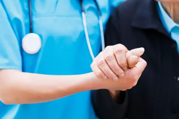 A healthcare worker in blue scrubs and a stethoscope helps an elderly person by holding their hand.