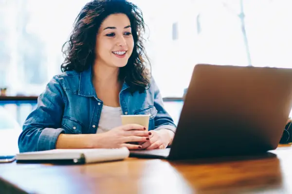 A person smiles while holding a cup and looking at a laptop screen, sitting at a table with a notebook.
