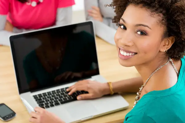 Person sitting at a table with a laptop, smiling at the camera. A smartphone is placed on the table nearby.