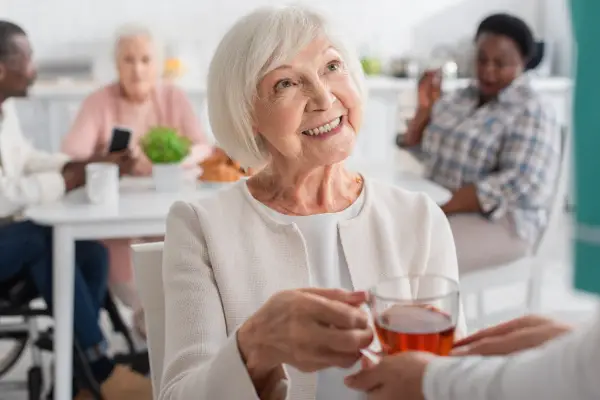 An elderly woman smiling holds a cup of tea offered by someone off-frame. In the background, people sit at a table engaged in conversation.