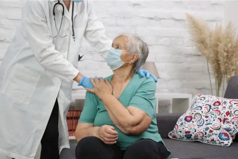 A doctor in a white coat and mask holds an elderly woman's shoulder as she sits on a couch, wearing a mask and teal top.