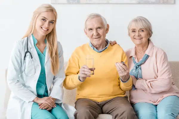 A doctor and an elderly couple sit on a sofa. The elderly man holds a glass of water and pills. The woman is smiling.