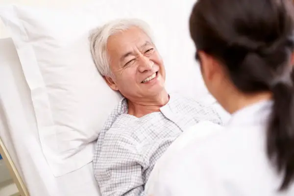 An elderly man smiles while lying in a hospital bed, interacting with a healthcare professional in a white coat.