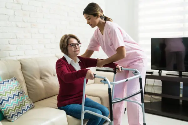 A caregiver in pink scrubs assists an elderly woman with glasses using a walker beside a couch in a living room.