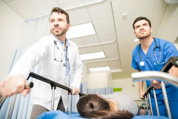 Two medical professionals, one in a white coat and the other in blue scrubs, are pushing a patient on a gurney through a hospital hallway.