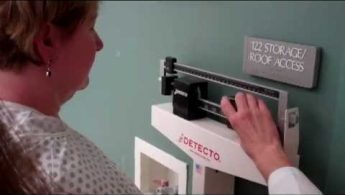 A person adjusts the weights on a beam balance scale near a green wall with a sign reading "122 Storage/Roof Access.